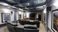 This single-floor private home theater is designed to deliver an immersive cinematic experience with refined architectural detail throughout. The room's most striking feature is its bold coffered ceiling, finished in deep navy and black tones, which lends a dramatic, upscale ambiance reminiscent of a classic movie palace. Recessed lighting embedded within the coffers provides warm, even illumination, while decorative columns with ornate capitals punctuate the walls and reinforce the room's formal character. Rows of plush black leather reclining chairs are arranged in a tiered layout, ensuring unobstructed sightlines to the large flat-screen display mounted at the front of the room. A built-in cabinetry unit beneath the screen provides storage and a polished finishing touch. A classic red popcorn machine adds a playful, authentic movie-house charm. Framed cinematic artwork adorns the walls, contributing to the room's dedicated theater identity. The two-tone wall treatment, featuring a deep wainscoting detail, complements the dark ceiling and enhances the immersive atmosphere. Carpeted flooring throughout adds acoustic comfort and a cozy feel, making this space an exceptional venue for film viewing and entertainment.