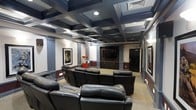 This single-floor private home theater is designed to deliver an immersive cinematic experience with refined architectural detail throughout. The room's most striking feature is its bold coffered ceiling, finished in deep navy and black tones, which lends a dramatic, upscale ambiance reminiscent of a classic movie palace. Recessed lighting embedded within the coffers provides warm, even illumination, while decorative columns with ornate capitals punctuate the walls and reinforce the room's formal character. Rows of plush black leather reclining chairs are arranged in a tiered layout, ensuring unobstructed sightlines to the large flat-screen display mounted at the front of the room. A built-in cabinetry unit beneath the screen provides storage and a polished finishing touch. A classic red popcorn machine adds a playful, authentic movie-house charm. Framed cinematic artwork adorns the walls, contributing to the room's dedicated theater identity. The two-tone wall treatment, featuring a deep wainscoting detail, complements the dark ceiling and enhances the immersive atmosphere. Carpeted flooring throughout adds acoustic comfort and a cozy feel, making this space an exceptional venue for film viewing and entertainment.