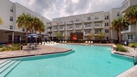 This expansive outdoor amenity space is designed to deliver a resort-like experience within a modern multi-story residential community. At its centerpiece sits a generously sized swimming pool with vivid turquoise water, framed by a paver deck and surrounded by rows of white lounge chairs ideal for sunbathing. A separate spa provides a dedicated relaxation zone, also lined with chaise lounges and anchored by tall palm trees that lend a distinctly tropical ambiance.

A standout feature of this outdoor space is a large-format LED screen positioned poolside, offering an outdoor entertainment experience for residents. Shaded dining and seating areas with market umbrellas provide options for those seeking respite from the sun.

The landscaping throughout is thoughtfully designed, incorporating native palms, ornamental grasses, and manicured greenery that soften the hardscape and create a lush, layered environment. Paver pathways wind through planted garden beds, connecting different zones of the courtyard seamlessly.

A dedicated lawn area with comfortable outdoor sofas and accent seating offers a casual gathering space, while raised stone planters add architectural dimension. The entire courtyard is enclosed by the surrounding multi-story building, creating an intimate and private retreat with a cohesive, contemporary aesthetic that balances relaxation, recreation, and community.