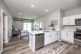 This thoughtfully designed single-floor residence offers a modern, open-concept layout that maximizes both functionality and comfort. The combined kitchen and living area serves as the heart of the home, featuring light wood-tone flooring, recessed lighting, and a large sliding glass door that floods the space with natural light and provides views of the outdoors. The kitchen is outfitted with white shaker-style cabinetry, a generous white quartz peninsula with bar seating, stainless steel appliances including a dishwasher and refrigerator, and an electric range—all accented by exposed industrial-style ductwork that adds a distinctive architectural character throughout the unit. The dedicated office provides a quiet retreat with plush carpeting, a large window overlooking the exterior, and ample room for a functional workspace. The primary bedroom offers a comfortable, carpeted retreat with a ceiling fan and a sizable window. The en-suite bathroom features a dual-sink white vanity with quartz countertops and direct access to a walk-in closet equipped with wire shelving systems. In-unit laundry is conveniently housed in its own dedicated room with full-size washer and dryer hookups and overhead shelving. The overall aesthetic is clean, bright, and contemporary throughout.