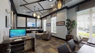 This well-appointed professional office space is thoughtfully designed to balance functionality with a sophisticated aesthetic. The main area serves as both a reception and lounge, featuring a coffered ceiling with dark trim accents and a pair of stylish globe pendant lights that anchor the space with a modern flair. Multi-pane windows fitted with slatted blinds and woven roman shades flood the interior with abundant natural light while offering views of lush greenery outside. The lounge area is furnished with plush upholstered armchairs arranged around a round wood-topped coffee table with an ornate metal base, creating a comfortable setting for informal meetings or waiting. A wall-mounted flat-screen television, built-in dark cabinetry, and a desktop computer workstation provide practical amenities for residents or staff. Framed artwork and a tall potted tree add warmth and visual interest throughout. A private meeting room, accessed through frosted glass double doors, offers an intimate setting with a rectangular table, sleek chrome-framed chairs, and decorative wall panels. The patterned carpet tile flooring in warm earth tones ties the entire space together, contributing to an environment that is both professional and inviting.