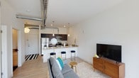 This single-floor, one-bedroom, one-bathroom residence offers a thoughtfully designed open-concept layout that maximizes its compact footprint. The living area flows seamlessly into the kitchen, creating a cohesive and functional space ideal for everyday living and entertaining. Light hardwood-style flooring runs throughout, contributing to a bright, airy atmosphere complemented by clean white walls.

A standout architectural detail is the exposed silver ductwork running along the ceiling, lending an industrial character that contrasts stylishly with the otherwise polished interior. The kitchen is well-appointed with dark cabinetry, stainless steel appliances including a refrigerator and microwave, and stone-look countertops. A peninsula with bar seating provides additional dining and workspace, highlighted by pendant lighting overhead.

The living area is open and inviting, with ample room for comfortable furnishings. Recessed lighting ensures consistent illumination throughout. The combination of modern industrial elements—exposed ductwork, pendant fixtures—with warm wood tones and neutral finishes creates a balanced, contemporary ambiance suited to a variety of tastes and lifestyles.