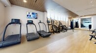 Located on Santa Monica Boulevard in Los Angeles, TEN(310) is a sleek, well-appointed fitness facility designed to deliver a premium workout experience. The space is thoughtfully designed with a clean, contemporary aesthetic, featuring warm wood-paneled ceilings, crisp white walls, and a combination of light hardwood flooring and durable fitness flooring that delineates training zones.

The main exercise room is generously proportioned and bathed in natural light from a row of clerestory windows along one wall, creating a bright and energizing atmosphere. The cardio zone is well-stocked with treadmills, stair climbers, ellipticals, stationary bikes, and rowing machines, all positioned for easy access and optimal spacing. A wall-mounted flat-screen television adds entertainment options during workouts.

The strength training area features a multi-functional cable and power rack system, a comprehensive dumbbell and kettlebell rack, weight plates, and a dedicated bench press station. Full-length mirrors enhance the space by amplifying light and allowing for form checks during exercise.

A welcoming reception and amenity area greets visitors at the entrance, complete with a marble-topped service counter, rolled towel storage, and access to restrooms. Glass doors with the TEN(310) branding provide an elegant transition into the main training floor, reinforcing the facility's polished, professional identity.