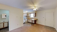 This single-floor apartment at Katelyn Court Apartments offers a practical and comfortable living experience with an open, functional layout. The living area features soft carpeting and a ceiling fan with integrated lighting, creating a cozy and well-ventilated atmosphere. Seamlessly connected to the living space, the kitchen is designed for everyday convenience and is equipped with white appliances, including a range and refrigerator. Dark wood cabinetry provides ample storage, while the light-toned countertops offer generous prep space. A peninsula counter with decorative spindle supports extends into the living area, serving as a natural divider between the two spaces and providing additional seating or serving space. The kitchen also benefits from a window above the sink, allowing natural light to brighten the workspace. The transition between the carpeted living area and the tile flooring of the kitchen creates a clear and practical delineation of the two zones. The neutral color palette throughout — featuring soft cream walls and light ceilings with a textured finish — gives the space a clean, understated aesthetic that is easy to personalize and maintain.