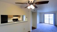 This single-floor residence offers a practical and comfortable layout designed for everyday living. The open-concept design connects the kitchen and living areas seamlessly, creating a sense of flow and spaciousness throughout. A defining feature of the main living area is the ceiling fan with an integrated light fixture, providing both air circulation and ambient lighting. The kitchen is accessible via a pass-through counter window, which opens to the living space and allows for easy interaction between rooms. The kitchen is equipped with a gas range and an overhead range hood, complemented by warm wood-toned cabinetry. The flooring transitions from hard surfaces near the kitchen to plush carpeting in the living and adjoining areas, adding warmth underfoot. A hallway visible from the main living space leads to additional rooms, hinting at a well-organized floor plan. Natural light filters in through windows and a glass door, brightening the interior. The textured ceiling and neutral wall tones throughout give the space a clean, classic aesthetic that is easy to personalize. Overall, this residence offers a straightforward and welcoming layout suited for comfortable daily living.