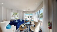 This single-floor community clubhouse is designed with a coastal aesthetic, combining functionality and leisure in a bright, welcoming environment. The main lounge area is bathed in natural light streaming through expansive floor-to-ceiling windows and glass doors that open directly to an outdoor pool area. Rich hardwood flooring runs throughout, complementing a crisp white and navy blue color palette accented by sheer floor-length curtains that add an airy, elegant touch. The lounge is furnished with a bold navy sofa, cream wingback chairs, rattan accent seating, and a geometric chandelier overhead, creating a sophisticated yet relaxed gathering space. An adjoining seating nook offers a quieter retreat with additional chairs and a console table. The game room is a highlight of the space, featuring a full-length shuffleboard table, a cozy TV lounge with blue velvet ottomans, and a large decorative Scrabble board on the wall. A dedicated business center lines one wall with a row of desktop computer workstations illuminated by pendant lighting. Throughout the space, recessed lighting and carefully chosen artwork contribute to a polished, resort-like atmosphere. Direct access to the pool deck further enhances the indoor-outdoor connectivity of this well-appointed community space.