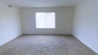 This single-story space presents a clean, minimalist interior designed to offer a blank canvas for personalization. The room visible in the image is finished with wall-to-wall neutral-toned carpeting, providing a soft, comfortable underfoot feel that complements the overall understated aesthetic. The walls are painted in a crisp, light neutral shade, contributing to the bright and airy atmosphere throughout the space.

A prominent feature of the room is the large window fitted with horizontal blinds, which allows an abundance of natural light to filter in, enhancing the sense of openness. The window serves as a focal point on the wall, drawing the eye and giving the room a welcoming, light-filled quality.

The layout appears straightforward and open, with clean corners and a smooth ceiling that reinforces the simple, functional design. The absence of decorative trim or ornate detailing gives the space a modern, no-frills character well-suited to a variety of furnishing styles. Overall, the space exudes a calm, neutral ambiance that provides a versatile foundation for comfortable everyday living.