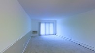 This single-floor space offers a clean and functional layout designed for comfortable everyday living. The main living area is notably open and spacious, featuring soft wall-to-wall carpeting that adds warmth underfoot. Large sliding glass doors dominate one wall, dressed with vertical blinds that allow for flexible light control while filling the room with an abundance of natural light. The bright, neutral-toned walls and white ceiling contribute to an airy, open ambiance throughout the space. A wall-mounted air conditioning unit provides climate control, complemented by a baseboard heating system that runs along the perimeter of the room, ensuring year-round comfort. The room's layout is versatile, providing ample space for a variety of furniture arrangements. The overall design is straightforward and practical, with clean lines and a minimal aesthetic that makes the space feel both welcoming and adaptable. The generous natural light from the large glass doors enhances the sense of openness, making this a bright and inviting environment suitable for a range of uses.