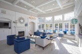Located at 980 Walther Blvd in Lawrenceville, Georgia, this single-story community clubhouse is designed with a refined, classical aesthetic that blends sophistication with comfort. The main lounge area impresses with soaring ceilings adorned with detailed coffered and crown molding, crisp white wainscoting panels, and wide-plank light hardwood flooring that runs throughout. A grand decorative fireplace serves as a focal point, framed by an ornate white surround and mantel, complemented by a stylish sunburst mirror above. Elegant arched doorways connect the various areas of the space, creating a seamless and inviting flow. Deep navy blue velvet seating and soft gray accent chairs are arranged atop patterned area rugs, adding warmth and a curated color palette to the interior. An abundance of oversized windows and French doors flood the space with natural light while offering direct views of the outdoor pool area, visually extending the interior. A service counter area, visible through one of the arched passages, provides additional functionality. Contemporary artwork in cool blue tones adorns the walls, reinforcing the cohesive design theme. The overall ambiance is bright, airy, and welcoming, combining classic architectural details with modern furnishings for an upscale communal experience.