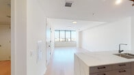 This modern, single-floor unit is designed with a clean, minimalist aesthetic throughout. Light-toned hardwood flooring flows seamlessly across the open living areas, complementing the crisp white walls and creating a bright, airy atmosphere.

The open-concept living and dining area is anchored by expansive floor-to-ceiling windows that frame sweeping panoramic city views, flooding the space with abundant natural light. The kitchen is a standout feature, showcasing a large waterfall-edge quartz island with an undermount sink and matte black fixtures. Sleek, glass-front cabinetry in a muted tone lines the walls above a full suite of stainless steel appliances, including a refrigerator, range, and microwave. Track lighting overhead adds both function and a contemporary touch.

The bedrooms continue the clean, modern palette with light wood flooring, white walls, and generous window exposure offering additional city vistas. Closet access is neatly integrated into the room design.

The bathroom is finished with large-format floor tiles and bold, dark wall tiles surrounding a soaking tub and shower combination. A floating wood-toned vanity with matte black fixtures adds warmth and contrast. The overall ambiance of this unit is polished, contemporary, and well-lit, ideal for those who appreciate modern urban living.