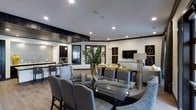 This single-floor communal space is thoughtfully designed with a modern aesthetic, seamlessly blending a dining area, lounge, and kitchenette into one open-plan layout. The dining area is anchored by a sleek glass-top table surrounded by upholstered gray chairs, offering an elegant setting for gatherings. Recessed lighting throughout the white ceiling illuminates the space evenly, enhancing the bright and welcoming atmosphere.

The lounge area features plush seating in neutral tones accented with gold and yellow cushions, arranged around a wall-mounted flat-screen television. Geometric wall art in black and gold adds a sophisticated decorative touch, complementing the contemporary design scheme. A tall tropical plant serves as a natural focal point between the dining and lounge zones, adding a refreshing organic element to the interior.

The kitchenette, visible in the background, is fitted with dark cabinetry, stainless steel appliances, and a light countertop, providing functional amenities for residents or guests. Dark wood trim details frame the transition between zones, lending depth and structure to the overall design. The space strikes a balance between functionality and upscale style.