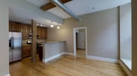 This single-floor space showcases a compelling blend of industrial and contemporary design elements. The open-concept layout connects the kitchen and living areas seamlessly, creating a spacious and airy feel enhanced by elevated ceilings. Exposed metal ductwork runs prominently overhead, complemented by rustic wooden support beams that add warmth and character to the industrial aesthetic.

The kitchen is equipped with warm-toned wood cabinetry, black appliances, and a granite-topped breakfast bar that serves as both a functional workspace and a natural divider between the cooking and living areas. Pendant lighting hangs above the bar, providing focused task lighting while contributing to the overall ambiance. Track lighting above the kitchen ensures the workspace is well-lit.

Light hardwood flooring extends throughout the space, tying the open areas together with a clean, cohesive look. Neutral gray walls provide a versatile backdrop that complements the mix of rustic and modern elements. A doorway visible from the main area leads to an additional room, suggesting flexible use of space. The overall atmosphere is modern yet characterful, ideal for those who appreciate loft-style living.