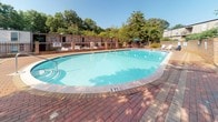 Located at 702 Edwards Rd in Greenville, South Carolina, this single-story outdoor amenity space offers a welcoming environment designed for relaxation and leisure. The centerpiece of the area is a large, kidney-shaped swimming pool with crystal-clear blue water, framed by a classic brick paver pool deck that provides ample space for lounging. Poolside seating, including lounge chairs and umbrellas, is available throughout the deck for comfort and shade.

Adjacent to the pool is a spacious brick paver patio area that extends toward the main building, which features a brick exterior and white-framed French doors that open directly to the outdoor space. The patio is furnished with wrought iron seating and umbrella-shaded tables, creating an inviting spot for outdoor dining or casual gatherings. Raised garden beds filled with dense, lush greenery and mature shrubs add a natural, landscaped feel to the surroundings.

The entire outdoor area is enclosed by iron fencing and surrounded by a canopy of mature trees, offering privacy and a shaded, park-like atmosphere. The combination of classic brick architecture, thoughtful landscaping, and well-appointed outdoor furnishings makes this space both functional and visually appealing — ideal for enjoying the warm Greenville climate.