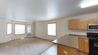 This single-floor space features an open-concept layout that connects the living area and kitchen, creating a functional and cohesive environment. The living area is finished with wall-to-wall carpeting and neutral-toned walls, lending a simple, understated aesthetic. Multiple windows with warm wood-toned frames allow natural light to filter throughout the space, contributing to a bright and airy atmosphere. A wall-mounted air conditioning unit and baseboard heating are visible, ensuring year-round climate comfort. The kitchen area adjoins the living space and is defined by a countertop overhang that serves as a natural divider between the two zones. Medium-toned wood cabinetry provides storage overhead, complemented by a dark laminate countertop. The overall design is straightforward and practical, with smooth, light-colored walls and a textured ceiling that add subtle visual depth. The open flow between the kitchen and living area makes the space feel generously proportioned, well-suited for everyday living and easy to furnish and personalize.