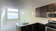 This single-floor space showcases a thoughtfully designed open-concept layout that connects the kitchen and living areas. The kitchen is a standout feature, equipped with sleek dark espresso cabinetry that provides a striking contrast against the light-toned countertops and walls. A kitchen island with an integrated stainless steel sink serves as a functional centerpiece, offering additional workspace and a natural gathering point. Two elegant pendant lights with frosted globe shades hang overhead, providing focused task lighting while contributing to the space's modern aesthetic. Additional upper cabinetry along the perimeter wall offers ample storage. The transition from the kitchen into the living area is seamless, creating an open and airy feel throughout. Large windows in the adjacent living area allow natural light to filter through, brightening the neutral-toned walls and light carpeted flooring. Recessed lighting supplements the natural light, ensuring the space remains well-lit throughout the day. The overall ambiance is clean, contemporary, and functional, with a neutral color palette that provides a versatile backdrop for a variety of interior styles.