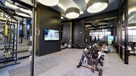 This contemporary fitness center is designed with both functionality and aesthetics in mind. The space is anchored by a dedicated cycling area, featuring a row of professional-grade stationary bikes with orange-accented details, ideal for high-intensity spin sessions. A wall-mounted flat-screen TV faces the cycling stations, allowing members to stay entertained or motivated during workouts. The interior design makes a bold statement with deep charcoal and black wall treatments that lend the space a sleek, upscale atmosphere. Large circular drum pendant lights are suspended from a tray ceiling, providing warm, ambient illumination that complements the modern décor. Full-length mirrors line the walls, visually expanding the space and allowing for proper form checks during exercise. Additional equipment, including a stability ball, is visible in the background, hinting at a broader range of workout options. The flooring consists of durable, low-pile carpet tiles suited for a fitness environment. Large windows along one wall invite natural light and offer views to the exterior, preventing the space from feeling enclosed. Overall, the fitness center combines a sophisticated design palette with practical, high-quality amenities to create an inviting and well-appointed workout environment.