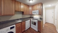 This single-story cottage offers a straightforward and functional layout designed for comfortable everyday living. The open living and dining area is generously sized, featuring warm wood-tone laminate flooring, crown molding, and large windows fitted with horizontal blinds that allow natural light to fill the space while providing views of the surrounding greenery outdoors.

The kitchen is efficiently arranged in an L-shaped configuration, showcasing warm honey-toned cabinetry with ample upper and lower storage, dark granite-style countertops, a stainless single-basin sink, and a full suite of white appliances including an electric range, built-in microwave, dishwasher, and refrigerator. A washer is conveniently located within the kitchen area, providing in-unit laundry access.

The bedroom is a comfortably proportioned room with soft carpeting, crown molding, a ceiling-mounted light fixture, a window overlooking greenery, and a reach-in closet with a hanging rod for storage.

The bathroom includes a light wood vanity cabinet, a white countertop, and wood-tone flooring consistent with the rest of the cottage. Overall, the space maintains a neutral, warm color palette throughout, creating a cohesive and inviting atmosphere.