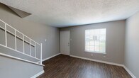This two-story home is designed with a practical and cohesive aesthetic throughout. The main level entry opens into a spacious living area finished with rich, dark hardwood-style flooring that extends across the lower floor. Neutral gray walls and white baseboards create a clean, understated palette, while a textured popcorn ceiling adds dimension overhead. A large window adjacent to the front door allows natural light to fill the living space, and a white-railed staircase leads to the upper level.

The kitchen is outfitted with white cabinetry, light countertops, and a full suite of white appliances, including a range, dishwasher, and refrigerator. A built-in shelving unit provides additional storage near the kitchen entry. The layout follows an efficient L-shape, maximizing counter and cabinet space.

Upstairs, the bedrooms feature soft carpeting, gray walls, and windows that bring in natural light. Ceiling fans with integrated lighting fixtures are installed for comfort. The bathrooms are finished with white vanity cabinetry, light stone-look countertops, large mirrors, and tub-shower combinations, offering functional and tidy spaces throughout.