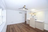 This single-floor unit offers a well-organized layout with a clean, neutral aesthetic throughout. Warm-toned hardwood flooring extends across the main living areas and bedrooms, lending a sense of continuity and warmth to the space. The open living and dining area is bright and airy, benefiting from multiple windows fitted with blinds that allow natural light to filter in, complemented by a ceiling fan overhead. A half-wall partition separates the living area from the kitchen, creating a sense of openness while maintaining distinct functional zones. The kitchen is outfitted with light wood cabinetry, a tile backsplash, a double sink, a dishwasher, and a full-size refrigerator, all set on clean white tile flooring. The bedrooms continue the hardwood flooring theme and are equipped with ceiling fans and a wall-mounted air conditioning unit for year-round comfort, with windows providing natural light and outdoor views. The bathroom features a pedestal sink, a toilet, and a full bathtub with a shower, accented by white mosaic tile walls and ample cabinet storage above. Overall, the space presents a practical and comfortable living environment with consistent finishes and functional amenities throughout.