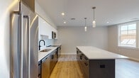 Located at 125 Warren St in Boston's Roxbury neighborhood, this single-floor residence showcases a thoughtfully designed modern interior with consistent finishes throughout. The open-concept kitchen and living area serves as the heart of the home, featuring a striking two-tone kitchen with high-gloss white upper cabinets, dark lower cabinetry, and a crisp white tile backsplash. A spacious center island with a white quartz countertop provides additional prep and seating space, while stainless steel appliances—including a French door refrigerator, range, microwave, and dishwasher—complete the chef-ready setup. Stylish cylindrical pendant lights hang above the island, adding a contemporary touch. The open living area extends generously from the kitchen, finished with light-toned hardwood-style flooring that runs throughout the entire unit. The bathrooms are elegantly appointed with white cabinetry, matte black hardware, quartz countertops, and large-format tile flooring. The primary bathroom features a dual-sink vanity and a separate walk-in shower with marble-style tile surround. Bedrooms are bright and well-proportioned, with neutral walls and natural light from windows offering neighborhood views. Recessed lighting is present throughout, contributing to a clean, modern ambiance.