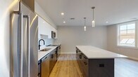 Located at 125 Warren St in Boston's Roxbury neighborhood, this single-floor residence showcases a thoughtfully designed modern interior with consistent finishes throughout. The open-concept kitchen and living area serves as the heart of the home, featuring a striking two-tone kitchen with high-gloss white upper cabinets, dark lower cabinetry, and a crisp white tile backsplash. A spacious center island with a white quartz countertop provides additional prep and seating space, while stainless steel appliances—including a French door refrigerator, range, microwave, and dishwasher—complete the chef-ready setup. Stylish cylindrical pendant lights hang above the island, adding a contemporary touch. The open living area extends generously from the kitchen, finished with light-toned hardwood-style flooring that runs throughout the entire unit. The bathrooms are elegantly appointed with white cabinetry, matte black hardware, quartz countertops, and large-format tile flooring. The primary bathroom features a dual-sink vanity and a separate walk-in shower with marble-style tile surround. Bedrooms are bright and well-proportioned, with neutral walls and natural light from windows offering neighborhood views. Recessed lighting is present throughout, contributing to a clean, modern ambiance.