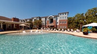 Located at 531 Old Maccumber Station Rd in Wilmington, NC, this outdoor amenity space presents a well-appointed, resort-style environment designed for relaxation and recreation. The centerpiece is a generously sized swimming pool with crystal-clear blue water, surrounded by a spacious sun deck lined with lounge chairs — ideal for enjoying North Carolina's sunny climate. A covered outdoor patio area provides a shaded retreat directly adjacent to the pool, furnished with comfortable seating including cushioned sofas and bar-height tables and chairs. A ceiling fan overhead ensures comfort during warmer months. The architecture throughout features a classic combination of warm red brick columns and accents paired with sage-green lap siding, lending the space a traditional yet refined character. Black iron fencing neatly encloses the pool area, enhancing both safety and aesthetic appeal. An outdoor-mounted television on the brick wall adds a modern entertainment element to the covered lounge. Mature trees surround the perimeter, providing natural shade and a lush backdrop. The multi-story residential buildings visible from the pool area reflect the same cohesive architectural style, creating a unified and inviting community atmosphere under open, sunlit skies.