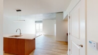 This single-floor space showcases a thoughtfully designed open-concept layout that seamlessly connects the kitchen, dining, and living areas. Light hardwood flooring runs throughout, complementing the clean white walls and contributing to a bright, cohesive aesthetic.

The kitchen is a standout feature, outfitted with rich warm-toned wood cabinetry extending to the upper walls, white quartz countertops, and a generous island with an undermount sink. Stainless steel appliances, including a refrigerator, range, and over-the-range microwave, are integrated alongside a subway tile backsplash and recessed and pendant lighting overhead.

The open living area benefits from an exposed concrete ceiling detail that adds an industrial character, contrasting elegantly with the otherwise clean, modern finishes. A sliding glass door leads to a private balcony with a metal railing, allowing abundant natural light to fill the space and offering views of the surrounding neighborhood.

The bathroom features light wood-grain flooring, a warm-toned wood vanity with a white countertop, an undermount sink, and a large framed mirror. Tiled walls in the shower area complement the overall neutral palette, creating a calm and functional space. The layout as a whole is efficient, well-lit, and designed with modern urban living in mind.