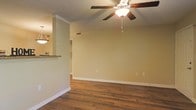 This single-floor space offers a clean, functional layout designed for comfortable everyday living. The main living area is highlighted by rich, dark hardwood flooring that runs throughout, adding warmth and a sense of continuity to the space. Crown molding trims the walls, lending a refined architectural detail that elevates the overall aesthetic. A ceiling fan with a built-in light fixture is centrally mounted, providing both air circulation and ambient lighting to the room.

The walls are finished in a soft, neutral tone that creates a calm and welcoming atmosphere, allowing natural light to reflect throughout the space. White baseboards provide a crisp contrast against the flooring, contributing to the tidy, well-maintained appearance of the interior.

Adjacent to the main living area, a partial wall with a countertop pass-through connects to the kitchen, where pendant lighting is visible overhead. This open-concept design promotes a natural flow between living and dining or kitchen areas, making the space feel connected and versatile. A textured ceiling adds subtle visual interest above. Overall, the space presents a practical yet inviting environment with classic interior finishes suited to a variety of uses.