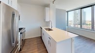 This single-floor unit is situated at an elevated position within a high-rise building, offering commanding views of the surrounding urban skyline through a dramatic wall of floor-to-ceiling windows. The expansive glazing floods the open living area with abundant natural light throughout the day, while hardwood-style plank flooring runs continuously across the main living space, contributing to a clean, cohesive aesthetic.

The kitchen is designed with functionality and style in mind. White flat-front cabinetry provides ample storage, complemented by light quartz countertops that extend into a peninsula with an integrated stainless steel sink. Stainless steel appliances — including a gas range, microwave, and refrigerator — complete the space. Track lighting overhead ensures practical illumination throughout.

The bathroom is finished to a high standard, featuring large-format marble-look porcelain floor tiles and matching wall tiles within the glass-enclosed walk-in shower. A wood-toned vanity with white countertop and backlit mirror adds warmth and contrast to the otherwise light, neutral palette.

Overall, the unit presents a modern, well-appointed interior with a strong emphasis on natural light, city views, and refined finishes throughout.