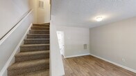 Located at 513 Verdoucq Dr in Holly Springs, NC, this two-story residence offers a practical and well-organized layout across two floors. The main level features wood-look flooring throughout the living room and kitchen areas. The living room receives natural light through a large window with views of surrounding greenery, and includes a wall-mounted HVAC vent for climate control. A textured ceiling and neutral wall tones carry throughout the home, creating a consistent, clean aesthetic. The kitchen is equipped with white cabinetry, a double-basin sink, electric range, and a full-size refrigerator, all finished in a cohesive white palette with light countertops and wood-look flooring. A carpeted staircase connects the two levels. Upstairs, both bedrooms are finished with plush carpet and feature windows that allow natural light to filter in. The primary bedroom includes two closets with hanging rods, offering ample storage. A secondary room with wood-look flooring provides flexible utility space. Both bathrooms feature a tub-shower combination, vanity with under-sink storage, and wood-look flooring, maintaining a clean and functional design throughout.