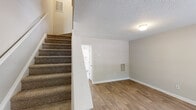 Located at 513 Verdoucq Dr in Holly Springs, NC, this two-story residence offers a practical and well-organized layout across two floors. The main level features wood-look flooring throughout the living room and kitchen areas. The living room receives natural light through a large window with views of surrounding greenery, and includes a wall-mounted HVAC vent for climate control. A textured ceiling and neutral wall tones carry throughout the home, creating a consistent, clean aesthetic. The kitchen is equipped with white cabinetry, a double-basin sink, electric range, and a full-size refrigerator, all finished in a cohesive white palette with light countertops and wood-look flooring. A carpeted staircase connects the two levels. Upstairs, both bedrooms are finished with plush carpet and feature windows that allow natural light to filter in. The primary bedroom includes two closets with hanging rods, offering ample storage. A secondary room with wood-look flooring provides flexible utility space. Both bathrooms feature a tub-shower combination, vanity with under-sink storage, and wood-look flooring, maintaining a clean and functional design throughout.