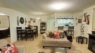 This single-floor residence offers a thoughtfully designed open-concept layout that brings together the main living areas in one cohesive and welcoming space. The living room serves as the heart of the home, featuring plush carpeted flooring and a comfortable seating area accented with decorative throw pillows and a patterned area rug that adds a touch of personality to the space. Adjacent to the living area, the dining space is anchored by a classic chandelier that casts a warm glow over the dark wood dining table and matching chairs, creating an intimate setting for meals and gatherings. A decorative shelving unit nearby adds both storage and visual interest to the room. The kitchen is accessible via a pass-through counter with bar-height seating, providing a convenient connection between the cooking and living spaces. White cabinetry lines the kitchen walls, complemented by a valance window treatment that introduces a charming, homey detail. A microwave and refrigerator are visible, rounding out the functional kitchen setup. Throughout the space, a textured ceiling adds a classic architectural detail, while the warm, neutral color palette and carefully chosen décor create an overall ambiance that is cozy, lived-in, and full of character.