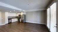 This single-floor space features an open-concept design that seamlessly integrates the kitchen and living area into one cohesive environment. The kitchen is outfitted with crisp white cabinetry, stainless steel appliances including a refrigerator, gas range, and built-in microwave, as well as a functional counter layout that maximizes workspace. Recessed lighting in the kitchen area provides focused illumination, complementing the natural ambient light throughout the space. The living area is generously proportioned, offering an open and flexible canvas for a variety of furniture arrangements. Dark hardwood-style flooring runs throughout the entire unit, providing a rich contrast against the neutral gray walls and bright white trim. The smooth white ceilings feature additional recessed lighting, contributing to a clean, contemporary aesthetic. A subtle step in the ceiling defines the transition between the kitchen and living zones, adding a mild architectural detail without interrupting the open feel of the layout. The overall design is modern and understated, emphasizing clean lines, functional finishes, and a neutral color palette well-suited to a variety of interior styles.