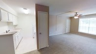 This single-floor residence offers a straightforward and functional layout designed for comfortable everyday living. The main living area is finished with wall-to-wall carpeting in a warm neutral tone, creating a cozy and cohesive atmosphere throughout the space. Walls are painted in soft, understated beige hues that contribute to a clean and inviting ambiance.

A notable architectural detail is the accent wall section rendered in a muted mauve or rose-toned paint, adding a subtle visual contrast to the otherwise neutral palette. This wall element also serves as a dividing feature between distinct areas of the space, helping to define zones without the use of full partitions.

The space includes a dedicated laundry area, visible with washer and dryer units, finished with light-toned tile flooring — a practical and durable surface choice for this utility zone. The transition between the tiled laundry area and the carpeted living space is clean and well-defined.

Ceiling-mounted light fixtures provide consistent illumination, while a ceiling fan in the living area supports air circulation. Multiple interior doors lead to additional rooms or storage areas, suggesting a well-organized floor plan. White trim and baseboards throughout lend a crisp, finished look that ties the interior together cohesively.