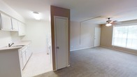 This single-floor residence offers a straightforward and functional layout designed for comfortable everyday living. The main living area is finished with wall-to-wall carpeting in a warm neutral tone, creating a cozy and cohesive atmosphere throughout the space. Walls are painted in soft, understated beige hues that contribute to a clean and inviting ambiance.

A notable architectural detail is the accent wall section rendered in a muted mauve or rose-toned paint, adding a subtle visual contrast to the otherwise neutral palette. This wall element also serves as a dividing feature between distinct areas of the space, helping to define zones without the use of full partitions.

The space includes a dedicated laundry area, visible with washer and dryer units, finished with light-toned tile flooring — a practical and durable surface choice for this utility zone. The transition between the tiled laundry area and the carpeted living space is clean and well-defined.

Ceiling-mounted light fixtures provide consistent illumination, while a ceiling fan in the living area supports air circulation. Multiple interior doors lead to additional rooms or storage areas, suggesting a well-organized floor plan. White trim and baseboards throughout lend a crisp, finished look that ties the interior together cohesively.