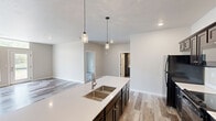 This single-floor residence offers a well-designed layout encompassing two bedrooms, two bathrooms, a combined kitchen and living area, two closets, and a dedicated laundry room. The open-concept kitchen and living space serves as the heart of the unit, featuring a large center island with a undermount double sink, white quartz-style countertops, dark cabinetry, black appliances including a refrigerator and range, and stylish pendant lighting overhead. Wood-look laminate flooring runs throughout the entire unit, providing a cohesive and contemporary aesthetic. The living area benefits from generous natural light via large windows and a glass-paneled entry door with transom window above, offering outdoor views of greenery. The primary bedroom is a well-proportioned space with a ceiling fan and a window, accompanied by an en-suite bathroom that includes a vanity with dark cabinetry and direct access to a walk-in closet outfitted with wire shelving systems. The second bedroom also features a ceiling fan, natural light, and outdoor views. The dedicated laundry room accommodates a full-size side-by-side washer and dryer and includes additional wire shelving for storage.