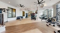 Located at 131 Great Circle Rd in Nashville, TN, this fitness center offers a thoughtfully designed, single-floor layout that balances functionality with modern aesthetics. Light wood-tone flooring runs throughout the space, complementing the crisp white walls and exposed brick accents that lend an industrial-chic character to the interior.

The main gym floor is generously sized and well-appointed, featuring a wide array of cardio equipment — including treadmills and elliptical machines — positioned in front of expansive floor-to-ceiling windows that flood the space with natural light and provide open views of the outdoor pool area and surrounding grounds. A free weights section lines the mirrored wall, complete with dumbbells, medicine balls, and resistance training equipment. Large ceiling fans enhance air circulation throughout the workout area.

A raised mezzanine platform at the center of the space, bordered by sleek black metal railings, provides visual definition and separation between workout zones. Adjacent to the gym floor, a lounge area offers a warm, inviting setting with a wood dining table, leather chairs, and a wall-mounted TV — ideal for relaxation or community gathering. Teal-painted doors add a distinctive pop of color, while restroom facilities are conveniently accessible. Recessed lighting throughout ensures a bright, welcoming environment at all times.