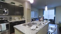 This single-floor space showcases a thoughtfully designed open-concept layout that seamlessly connects the kitchen and living area. The kitchen is a standout feature, boasting expansive granite countertops with subtle speckled detailing, a deep stainless steel sink, and sleek dark espresso cabinetry equipped with contemporary hardware. A subway tile backsplash adds a clean, classic touch, while modern pendant lighting illuminates the generous island workspace below. Upper cabinet space extends toward the ceiling, providing ample storage and drawing the eye upward to emphasize the room's vertical scale. The transition into the living area is effortless, where natural light pours in through large windows dressed with horizontal blinds and blue curtain panels. The living space is anchored by framed wall art and accented with blue throw pillows that add a pop of color against neutral-toned furnishings. The overall ambiance is modern and welcoming, with a cohesive color palette of warm grays, deep browns, and cool blues that lends a polished, contemporary feel throughout. The open flow between the kitchen and living room makes the space feel expansive and well-suited for both everyday living and entertaining.