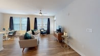 This single-floor residence presents a thoughtfully designed layout with modern finishes and a cohesive contemporary aesthetic throughout. The open living area is bright and welcoming, featuring warm hardwood-style flooring, a ceiling fan, and multiple windows dressed with navy drapes and white blinds that allow generous natural light to fill the space. The living room comfortably accommodates seating and entertainment areas, with direct access to an entry door framed by transom windows.

The kitchen is well-equipped with dark espresso cabinetry extending to the ceiling, light granite countertops, a blue-gray tile backsplash, and stainless steel appliances including a gas range, over-the-range microwave, and refrigerator. A small office or flex space features hardwood-style flooring and a sleek white desk with chrome legs.

The primary bedroom is carpeted and serene, featuring natural light from a window with blinds and abstract wall art above the bed. A secondary bedroom with ceiling fan offers flexibility as a guest room or multifunctional space. The bathroom includes an elongated dark vanity with granite countertops, a wide frameless mirror, and tile flooring, maintaining the refined material palette found throughout the residence.
