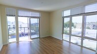 This single-floor space is defined by its open layout and an abundance of natural light streaming in through large sliding glass doors and oversized windows fitted with horizontal blinds. The living area features warm hardwood flooring that runs throughout, complementing the neutral, beige-toned walls and smooth white ceilings. Two sets of sliding glass doors provide direct access to separate outdoor balcony areas, each offering views of the surrounding urban and natural landscape, including tree canopies and neighboring structures. The dual balcony configuration is a standout architectural feature, effectively extending the living area outdoors while filling the interior with generous cross-light throughout the day. The corner positioning of the room allows for a spacious, airy feel, with walls meeting at an angle that enhances the sense of openness. The space is clean and unfurnished, making its structural features immediately apparent, including baseboards, recessed wall outlets, and a ceiling fixture point. The overall ambiance is bright, modern, and versatile, well-suited for a variety of furniture arrangements and living styles.