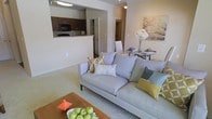 This single-story residence showcases an open-concept layout that connects the living room, dining area, and kitchen into one cohesive and functional space. The living room is carpeted and anchored by a contemporary light gray sofa accented with decorative pillows in muted yellows, greens, and teals, complemented by a warm wood-toned coffee table. A compact dining area sits adjacent, featuring a round glass-top table with white chairs, ideal for intimate gatherings. The kitchen is visible through a pass-through opening framed by a raised breakfast bar with granite-style countertops, offering both a casual seating option and a visual connection between cooking and living spaces. Dark espresso cabinetry lines the kitchen walls, paired with black appliances including a built-in microwave, contributing a bold contrast to the neutral, warm-toned walls throughout. Crown molding adds a refined architectural detail to the living areas. A hallway visible from the main space suggests additional rooms beyond, while the overall color palette of beige, taupe, and soft white creates a calm, welcoming ambiance throughout.