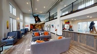 This expansive single-floor community clubhouse makes a striking first impression with its soaring double-height ceilings, exposed industrial ductwork, and an upper mezzanine level framed by sleek black metal railings. The grand lobby area centers around a rustic wood-paneled reception desk, flanked by plush gray sofas accented with bold orange and cowhide throw pillows — a nod to the space's nature-inspired design theme. A large-scale black bear mural dominates one wall, setting a vibrant, wilderness-themed aesthetic carried throughout via colorful wildlife paintings and sculptural décor pieces.

Wide-plank hardwood-style flooring flows seamlessly through the lounge, dining, and gathering zones. Floor-to-ceiling windows and glass sliding doors flood the interior with natural light and offer direct views of the outdoor pool and patio area, creating a strong indoor-outdoor connection. The dining area features round pedestal tables with upholstered chairs, ideal for casual gatherings. A private conference room, furnished with a large table and nature-themed artwork, provides a more intimate meeting space. A secure package and storage room offers practical functionality. Throughout, the space balances rustic, nature-inspired character with modern, open-plan design.
