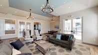 This single-story communal space is thoughtfully designed to foster comfort and social engagement. The main lounge area is anchored by soaring vaulted ceilings accented with a distinctive sky-blue tray ceiling detail, from which three wrought-iron chandeliers with candle-style lighting descend, adding a touch of elegance. The seating arrangement features plush gray sofas and wingback chairs adorned with teal and navy accent pillows, all arranged atop a patterned area rug over light hardwood flooring. A rustic wood coffee table serves as a central focal point.

A sleek, dark-tiled fireplace occupies one wall, providing a warm, inviting atmosphere. Large windows and glass-panel exterior doors flood the space with natural light while offering views of the surrounding outdoor area. Adjacent to the main lounge, a dedicated game room features a round poker-style table with leather chairs beneath a modern chandelier, set on a matching patterned rug. French-style interior doors connect the zones, maintaining visual openness throughout. The overall palette of warm beige tones, soft blues, and natural wood accents creates a cohesive, welcoming ambiance.