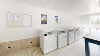 Located at 45 Barlake Avenue in Hamilton, Ontario, this shared laundry facility offers a clean, functional, and well-organized space designed to accommodate the needs of multiple users efficiently. The room is finished with crisp white walls and classic small-format mosaic tile flooring, lending it a tidy, practical character throughout.

The facility is equipped with a row of front-loading washing machines and a separate bank of top-vented dryers, all positioned along the walls for easy access. A large central folding table with a white surface and blue-painted legs provides ample workspace for sorting and folding laundry. Informational and instructional signage is posted on the walls, offering guidance on machine use.

Natural light filters generously into the space through large windows, which also provide views of the surrounding neighbourhood. Overhead fluorescent lighting ensures the space remains well-lit at all times. A utility sink is available near the entrance, adding to the room's functionality. The space also includes a bench along the window wall, offering a convenient place to wait.

Safety features include a visible fire extinguisher and no-smoking signage. Access is provided through a solid door, and a storage area is situated near the entry. Overall, this laundry facility is a well-maintained, practical communal space designed to serve its users effectively.