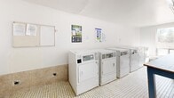 Located at 45 Barlake Avenue in Hamilton, Ontario, this shared laundry facility offers a clean, functional, and well-organized space designed to accommodate the needs of multiple users efficiently. The room is finished with crisp white walls and classic small-format mosaic tile flooring, lending it a tidy, practical character throughout.

The facility is equipped with a row of front-loading washing machines and a separate bank of top-vented dryers, all positioned along the walls for easy access. A large central folding table with a white surface and blue-painted legs provides ample workspace for sorting and folding laundry. Informational and instructional signage is posted on the walls, offering guidance on machine use.

Natural light filters generously into the space through large windows, which also provide views of the surrounding neighbourhood. Overhead fluorescent lighting ensures the space remains well-lit at all times. A utility sink is available near the entrance, adding to the room's functionality. The space also includes a bench along the window wall, offering a convenient place to wait.

Safety features include a visible fire extinguisher and no-smoking signage. Access is provided through a solid door, and a storage area is situated near the entry. Overall, this laundry facility is a well-maintained, practical communal space designed to serve its users effectively.
