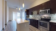 This single-floor residence showcases a contemporary design with an open-concept layout that seamlessly connects the kitchen and living areas. The kitchen is a standout feature, equipped with full stainless steel appliances including a range, microwave, and dishwasher, complemented by rich dark cabinetry that extends to the ceiling for ample storage. A multicolored tile mosaic backsplash adds visual texture, while light countertops provide contrast. A center island with a smooth, light-toned surface offers additional workspace and casual seating. Pendant lighting hangs above, and wood-look flooring runs throughout the main living areas, adding warmth to the modern aesthetic. A ceiling fan is positioned over the living area, which benefits from natural light through windows and a sliding glass door opening to a private balcony. The bathroom features large-format white tile flooring, a soaking tub with a glass enclosure, and a dark-cabinetry vanity with a mirror. Adjacent to the bathroom, a generously sized walk-in closet is outfitted with multi-level wire shelving. The bedroom is carpeted and receives natural light through a window with blinds, offering a quiet and comfortable retreat.