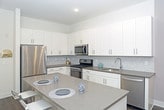 This single-floor space showcases a contemporary open-concept kitchen designed with both functionality and style in mind. The kitchen is anchored by a generously sized central island topped with a smooth quartz countertop in a warm gray tone, offering ample prep space and casual seating for two. Crisp white shaker-style cabinetry lines the walls, providing abundant storage, while a stainless steel refrigerator adds a polished, modern touch. A subtle mosaic tile backsplash behind the cabinetry introduces a delicate textural element without disrupting the clean aesthetic. Dark hardwood-style flooring runs throughout, creating a striking contrast against the white cabinetry and light walls. Recessed lighting illuminates the space efficiently, contributing to a bright and airy atmosphere. The neutral white walls are accented with a framed piece of artwork, adding a touch of warmth and personality to the otherwise minimalist design. The overall ambiance is clean, modern, and inviting, with a cohesive color palette of whites, grays, and dark wood tones that lend the space a polished, move-in-ready appeal.