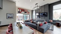 This single-story clubhouse presents a thoughtfully designed communal space that blends modern aesthetics with functional comfort. The main lounge area features soaring ceilings that amplify the sense of openness, anchored by a striking geometric pendant light fixture that serves as a focal point overhead. A large-scale abstract artwork in red, white, and black tones adds a bold visual statement to the neutral wall palette.

The lounge is furnished with a generously sized sectional sofa in charcoal gray, accented with red and white throw pillows that echo the room's color scheme. A wall-mounted flat-screen television is set against a dramatic dark paneled accent wall composed of textured square modules, creating a sophisticated backdrop. A concrete-style communal table with blush-toned seating occupies the foreground, offering a casual gathering or dining area.

Large sliding glass doors draw natural light deep into the interior while providing direct access to an outdoor patio space, where additional seating is visible. The herringbone-patterned hardwood flooring adds warmth and texture, tying together the contemporary design elements throughout the space.