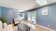 Located at 200 Mal Mae Ct in Williamsburg, Virginia, this pool house is a single-floor structure designed for comfort and convenience. The interior is finished with light hardwood flooring throughout, complementing the calming blue-gray walls and crisp white trim and baseboards that define the space's clean, coastal aesthetic.

The main lounge area is bright and open, enhanced by a large skylight that floods the room with natural light. Recessed lighting provides additional illumination. The lounge is equipped with comfortable seating — including a sofa and swivel chairs arranged around a wood-slat coffee table — as well as a wall-mounted flat-screen television, making it an ideal space for relaxation or entertainment. A patterned area rug anchors the seating arrangement over the hardwood floor.

Large French doors open directly to the outdoor pool area, offering unobstructed views and easy access. Adjacent windows further frame the poolside scenery and enhance the connection to the outdoors.

A functional kitchenette is tucked along one wall, outfitted with white cabinetry, granite countertops, stainless steel appliances including a full-size refrigerator and microwave, and a utility sink. A bathroom rounds out the layout, providing full convenience for pool guests. This space is thoughtfully designed to serve as a complete, self-contained amenity.