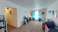 This single-floor residential space offers a comfortable and functional layout designed for everyday living. The living room serves as the heart of the home, featuring wall-to-wall carpeting that adds warmth underfoot and a classic textured ceiling overhead. A large window dressed with vibrant teal curtains layered over sheer white panels allows natural light to fill the room while adding a pop of color to the neutral surroundings. The walls are painted in a clean, crisp white, providing a bright and airy backdrop that complements the space's cozy character. Decorative wall accents add a personal touch, contributing to the inviting ambiance. The room comfortably accommodates a seating area and an entertainment setup, with ample open floor space that enhances the sense of roominess. A hallway visible from the living area connects to additional rooms, suggesting a thoughtfully arranged floor plan. The overall atmosphere is warm and homey, making it a practical and comfortable space suited for relaxed, day-to-day living.